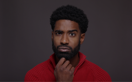 Homme avec barbe et cheveux crépus, portant un pull rouge, avec une expression pensive, se tenant le menton.