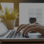 Une personne tape sur un clavier d'ordinateur portable, avec une plante en arrière-plan et une tasse posée près de l'écran.