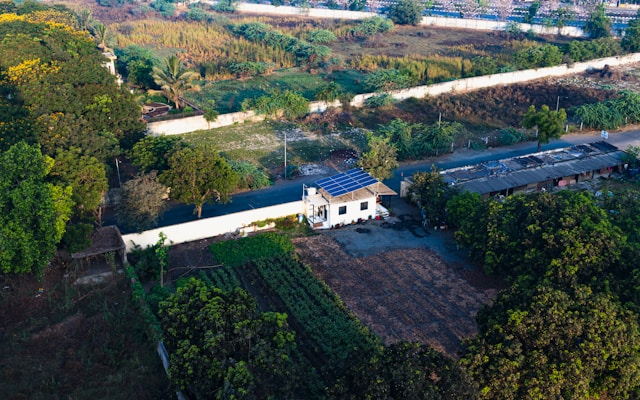 Vue aérienne d'une maison avec des panneaux solaires, entourée de champs et de végétation dans un environnement rural.