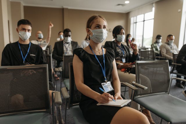 Participants masqués en conférence, une femme prend des notes au premier plan, d'autres écoutent attentivement ou posent des questions.