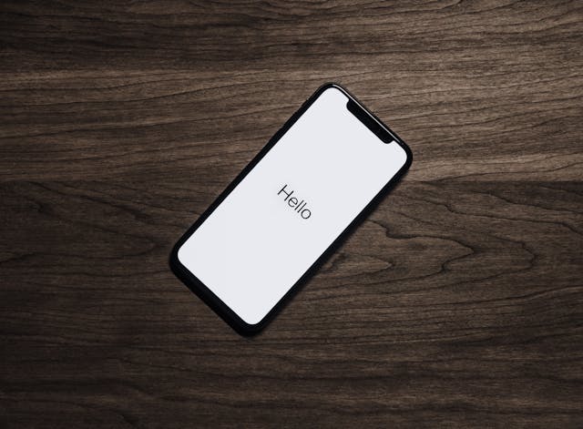 Un smartphone noir sur bois foncé affiche "Hello" en noir sur fond blanc, offrant une présentation minimaliste et évoquant un message de bienvenue.