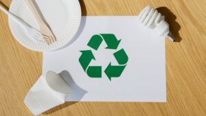Symbole de recyclage vert sur une feuille blanche, entouré d'objets recyclables : assiette en papier, couverts en bois, gobelet écrasé et ampoule.