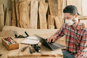 Un artisan en chemise à carreaux et masque de protection utilise un ordinateur dans un atelier, entouré d'outils et de planches de bois brut.