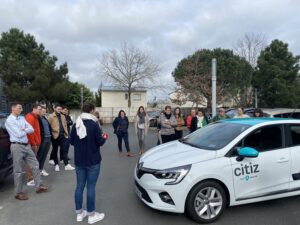 Des personnes en tenue décontractée entourent une voiture blanche marquée "Citiz local & national" sur un parking, attentives à une démonstration, avec arbres et bâtiments résidentiels en arrière-plan, reflétant une ambiance locale et conviviale.