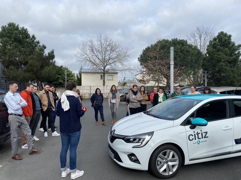 Des personnes en tenue décontractée entourent une voiture blanche marquée "Citiz local & national" sur un parking, attentives à une démonstration, avec arbres et bâtiments résidentiels en arrière-plan, reflétant une ambiance locale et conviviale.