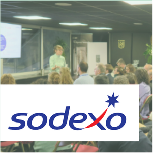 Un conférencier s'adresse à un public attentif dans une salle lumineuse avec sièges rouges, écran de projection, et le logo "Sodexo" en avant-plan.