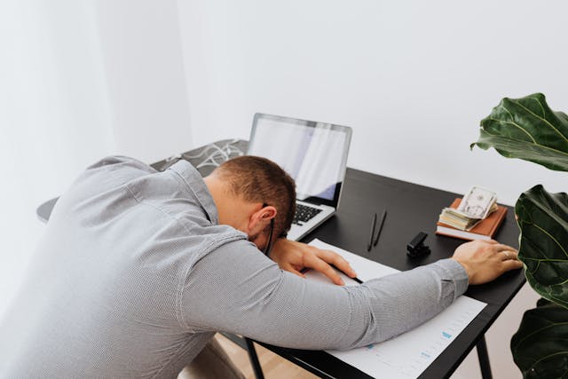Un homme en chemise grise, affalé sur un bureau noir avec ordinateur et livres, illustre fatigue dans un cadre épuré et moderne.
