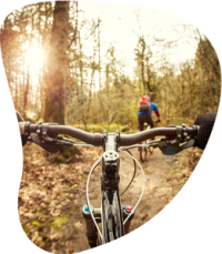 Créations Canva VRAC (18) Vue depuis un guidon de VTT sur un sentier forestier ensoleillé, avec un cycliste en arrière-plan, évoquant sport et nature.